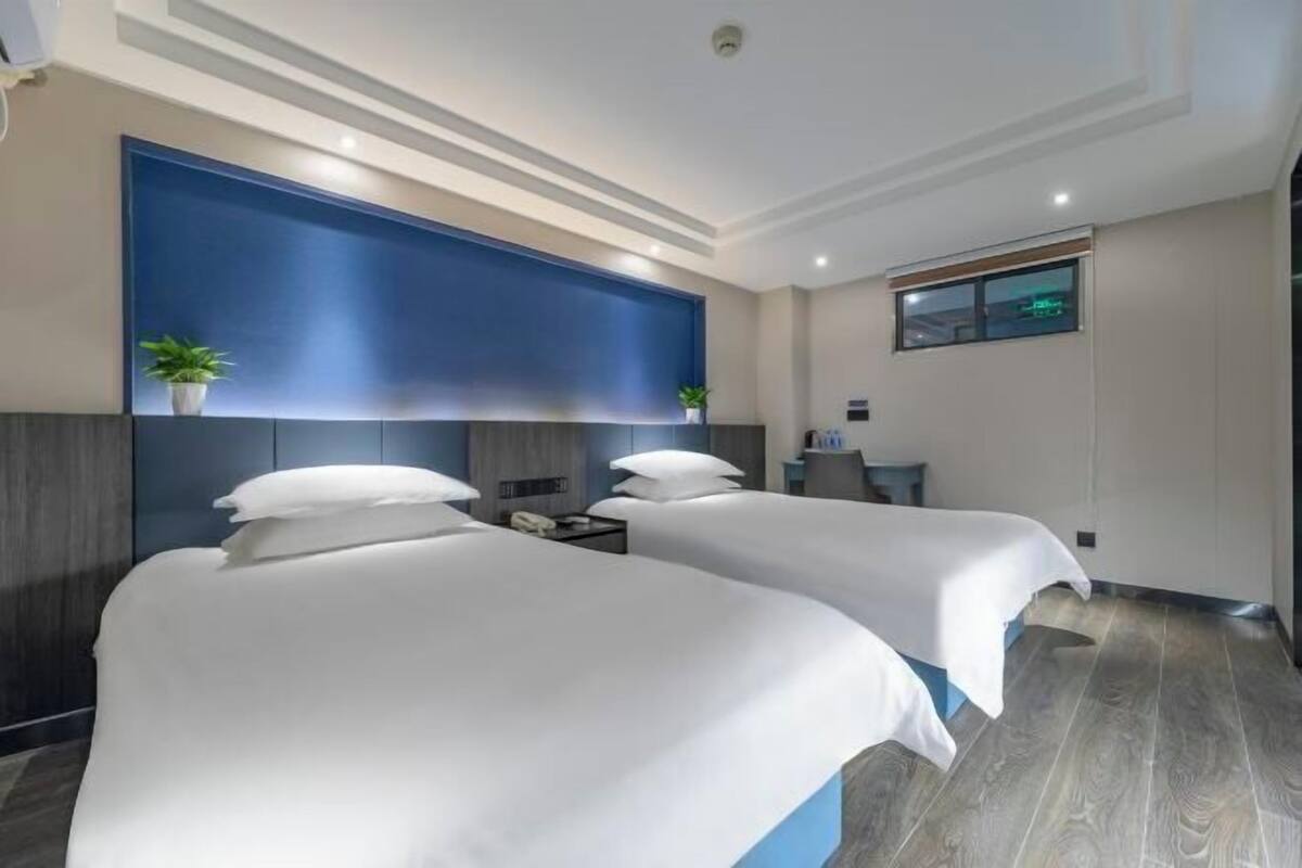 Ein Hotelzimmer mit zwei Betten, einer blauen Akzentwand, einem kleinen Fenster und einem Nachttisch.