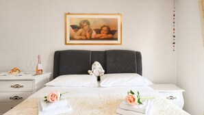 Un letto rifatto con un copriletto bianco e due rose rosa, un quadro con angioletti sopra il letto e un piccolo peluche sui cuscini.