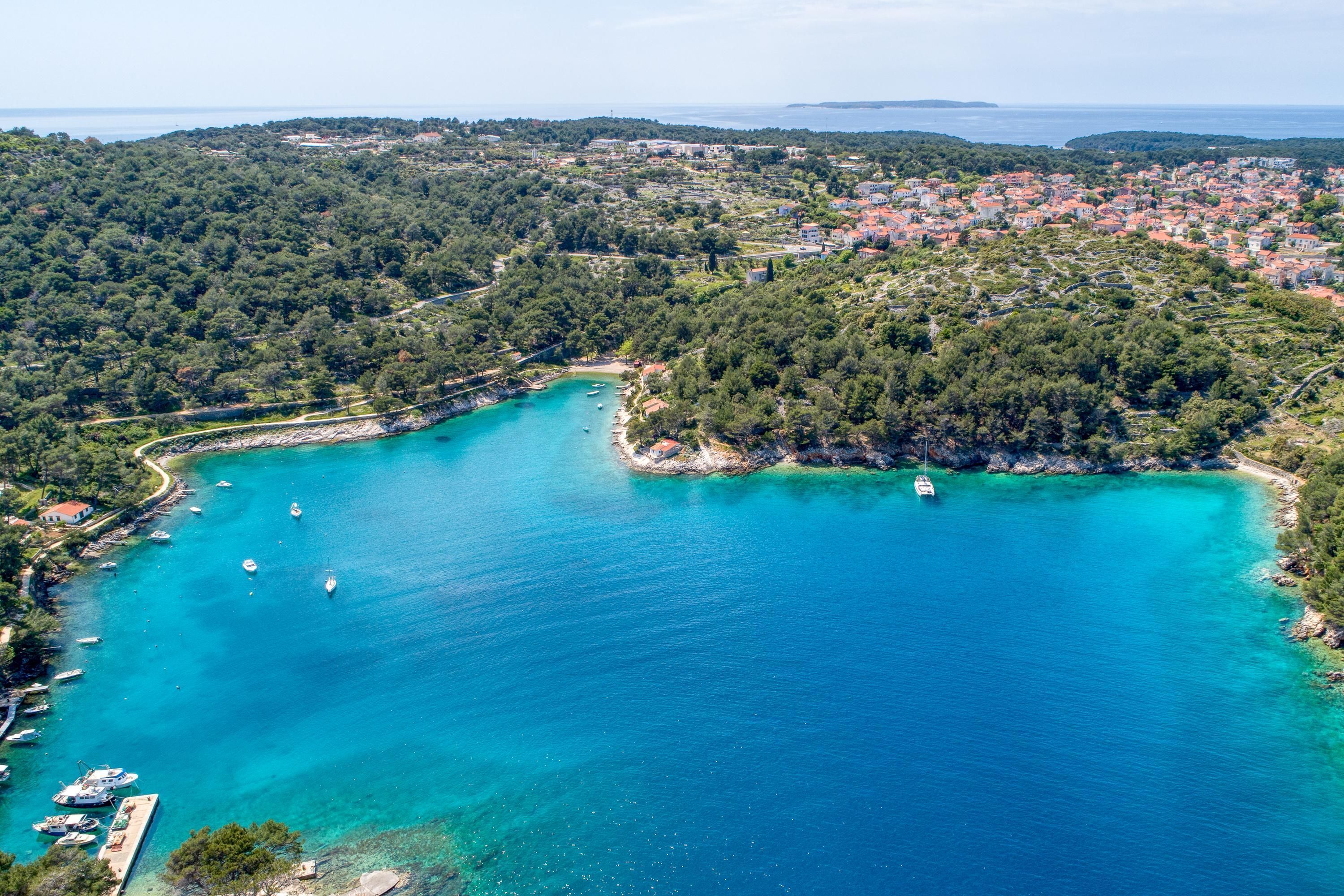 Vista aérea de uma vila costeira com uma baía turquesa, barcos e vegetação densa.