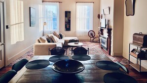A cozy living room with a wooden ceiling, a sofa, a coffee table, and a dining table with chairs. There are two windows with curtains, a mirror, and a shelf with decorative items. - Large, 2 BR stable conversion (circa 1874) tucked in the heart of old town. 🍃 (Echuca)
