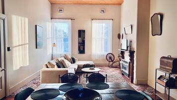 Un salon chaleureux avec un plafond en bois, un canapé, une table basse et une table à manger avec des chaises. Il y a deux fenêtres avec des rideaux, un miroir et une étagère avec des objets décoratifs.