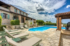 A poolside area with lounge chairs, a stone pool, and a stone building with green shutters.
