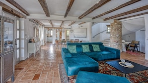 Un vaste espace de vie agrémenté d’un grand canapé d’angle turquoise, d’une table basse et d’un tapis à motifs. La pièce est dotée de poutres apparentes, d’un pilier en briques et offre une vue sur l’espace repas.