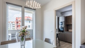 Eine moderne Küche mit Edelstahlgeräten, eine Essecke mit Glastisch und Stühlen sowie ein Blick auf einen Balkon mit Blumen.