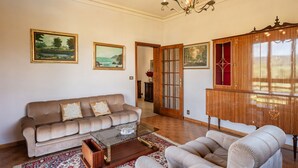 Een zithoek met een bank, fauteuil en een houten kast. Er hangen schilderijen aan de muur en er is een kroonluchter.
