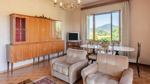 Een ruime woonkamer met een groot houten kastje, twee beige fauteuils, een televisie en een eettafel met stoelen. Een groot raam biedt uitzicht op een bergachtig landschap.