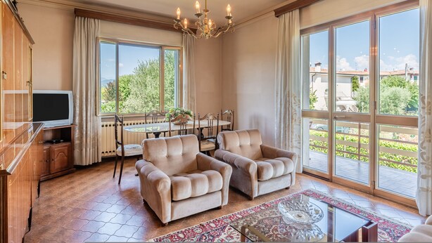 Een ruime woonkamer met twee beige fauteuils, een glazen salontafel en een kroonluchter. Een televisie is aan de muur bevestigd en er is een eethoek met een tafel en stoelen. Grote ramen bieden uitzicht op het omliggende landschap.
