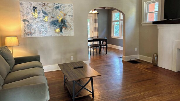 A living room with a sofa, a coffee table, a painting, a dining area with chairs, a fireplace, and a TV. - Palmetto 2 Bed Family House in Cincy near Oakley (Cincinnati)