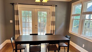 A dining room with a large wooden table and six chairs, a set of double doors with curtains, and a window with blinds. - Palmetto 2 Bed Family House in Cincy near Oakley (Cincinnati)