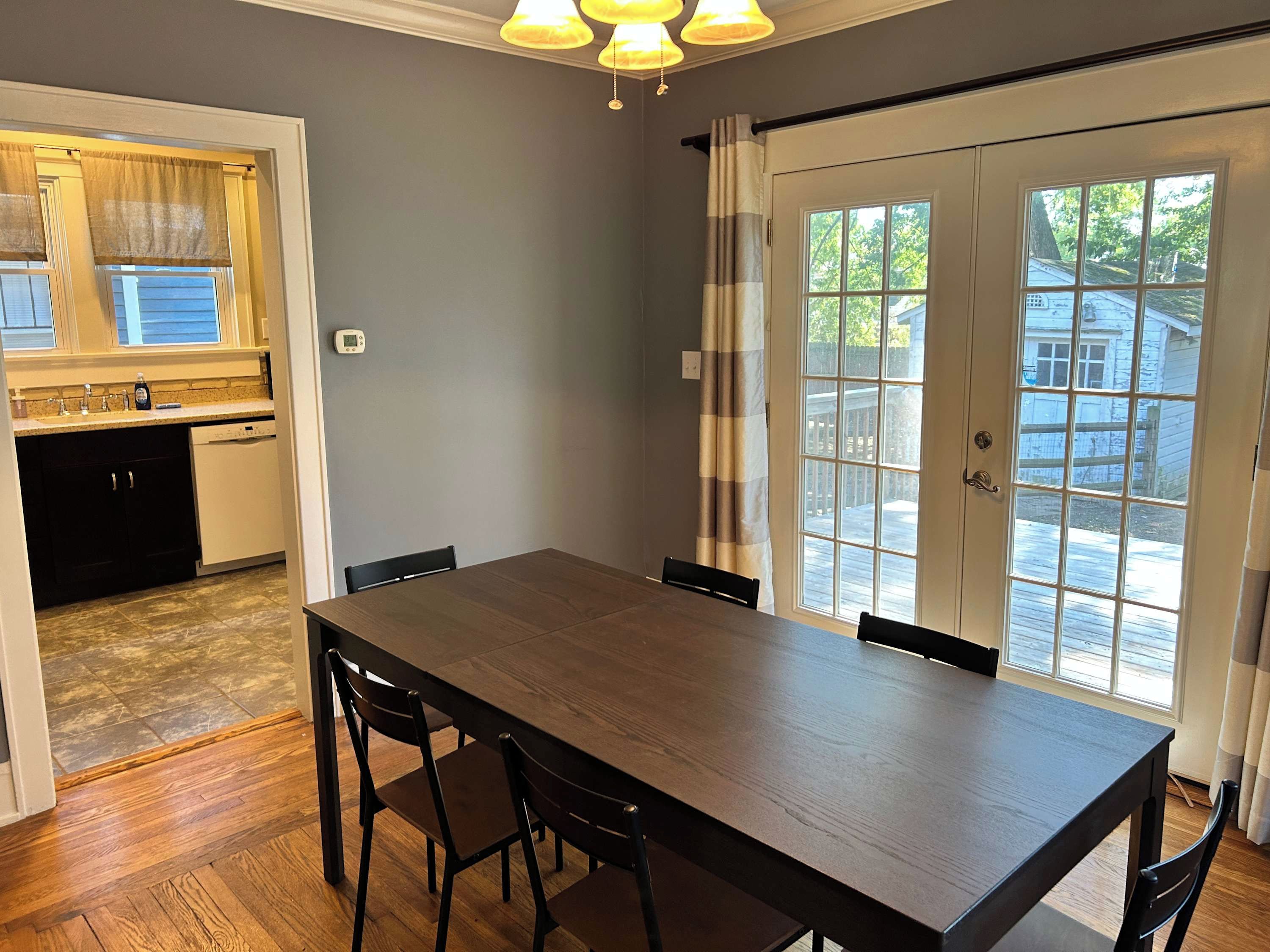 A dining area with a wooden table and chairs, a kitchen with a dishwasher, and a view through a large window with curtains.