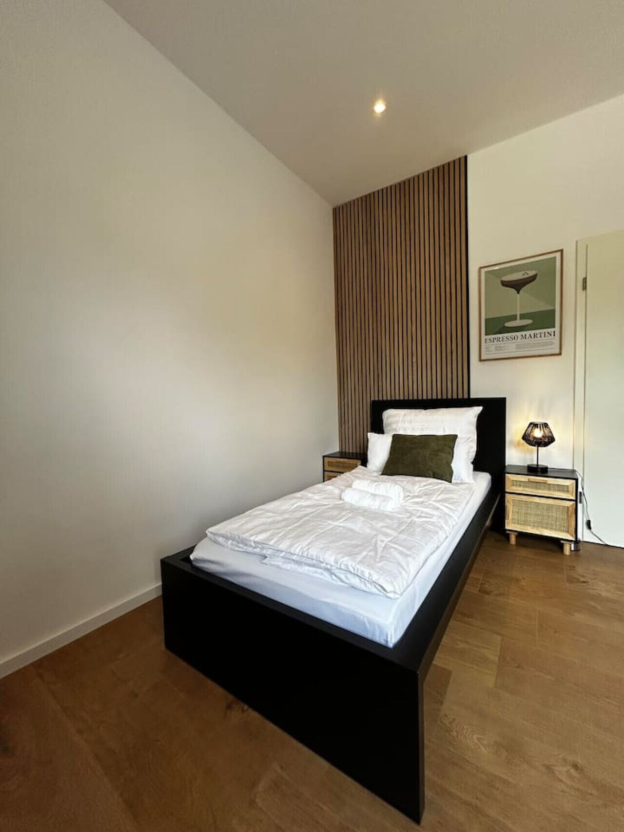 A modern bedroom with a black bed frame, white bedding, a wooden panel wall, a framed picture, and a bedside lamp.