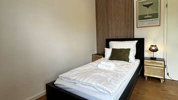 A modern bedroom with a black bed frame, white bedding, a wooden panel wall, a framed picture, and a bedside lamp.