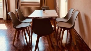 Een kamer met een tafel, vier stoelen, een raam met gordijnen, een radiator en een wandlamp.