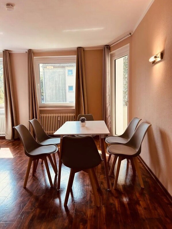 A room with a table and four chairs, a window with curtains, a radiator, and a wall-mounted light fixture.