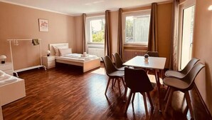 Een kamer met een bed, nachtkastje en een klein tafeltje met stoelen. Er is een raam met gordijnen, een radiator en een foto aan de muur.