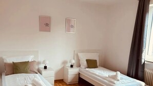 Habitación de hotel con dos camas individuales, suelo de madera, ventana con cortinas y dos mesitas de noche con lámparas.