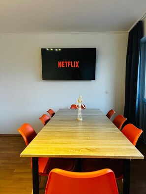 Een vergaderruimte met een lange tafel, zes oranje stoelen, een houten etalagepop en een televisiescherm waarop 'NETFLIX' staat.