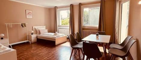 A room with a bed, bedside table, and a small table with chairs. There is a window with curtains, a radiator, and a picture on the wall.