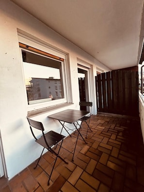 Un balcón con una mesa, dos sillas, una ventana y vistas a edificios.