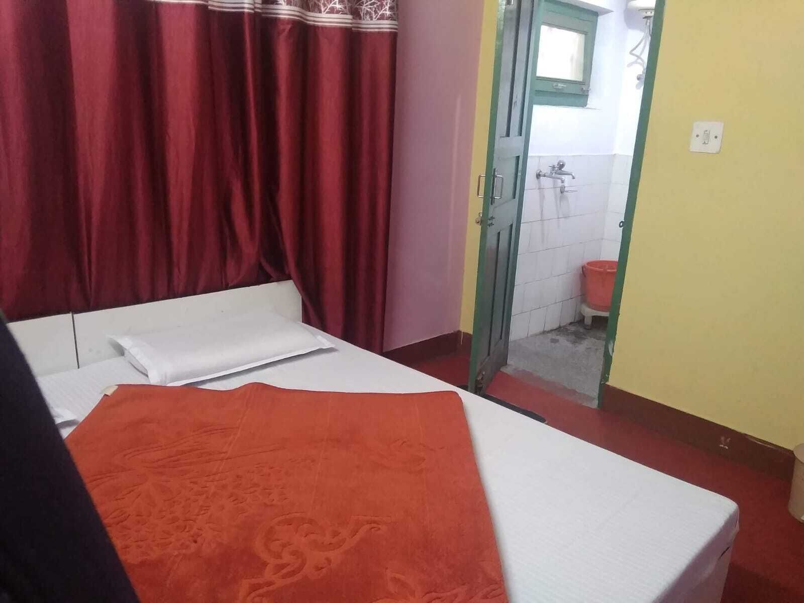Una habitación pequeña con cama individual, cortina roja, cubrecama blanco y manta roja.