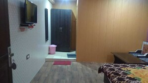A bedroom with a bed, a TV mounted on the wall, a wooden wardrobe, and a pink trash bin. - Hotel Grand Kailash (Joshimath)