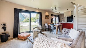 Ein modernes Wohnzimmer mit einer Couch, einem Hocker und Sesseln, ein Essbereich mit Tisch und Stühlen sowie eine Küche mit roten Schränken.