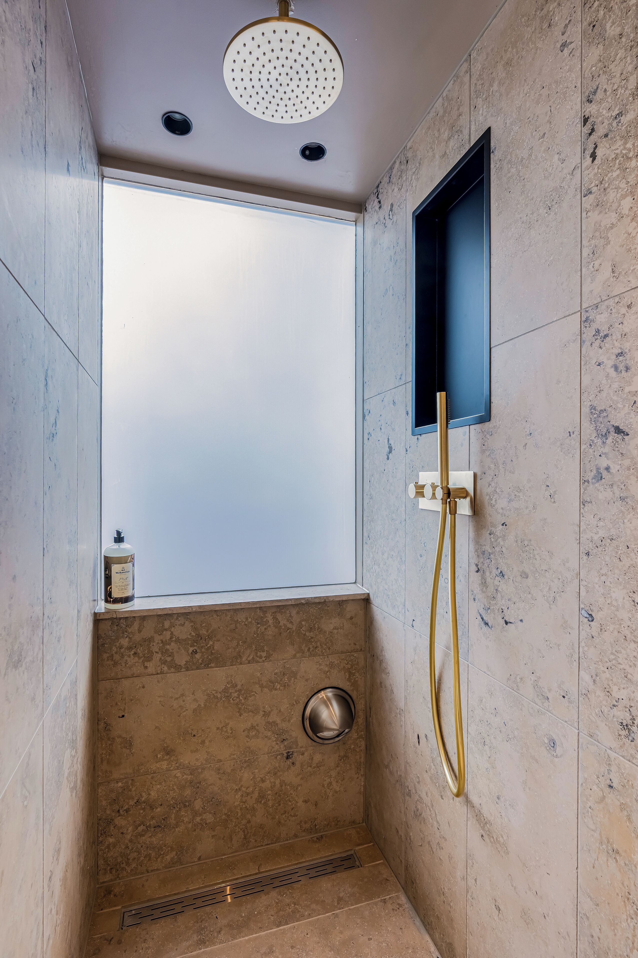 A shower with a glass door, a handheld showerhead, and a small bottle of shampoo on the ledge.