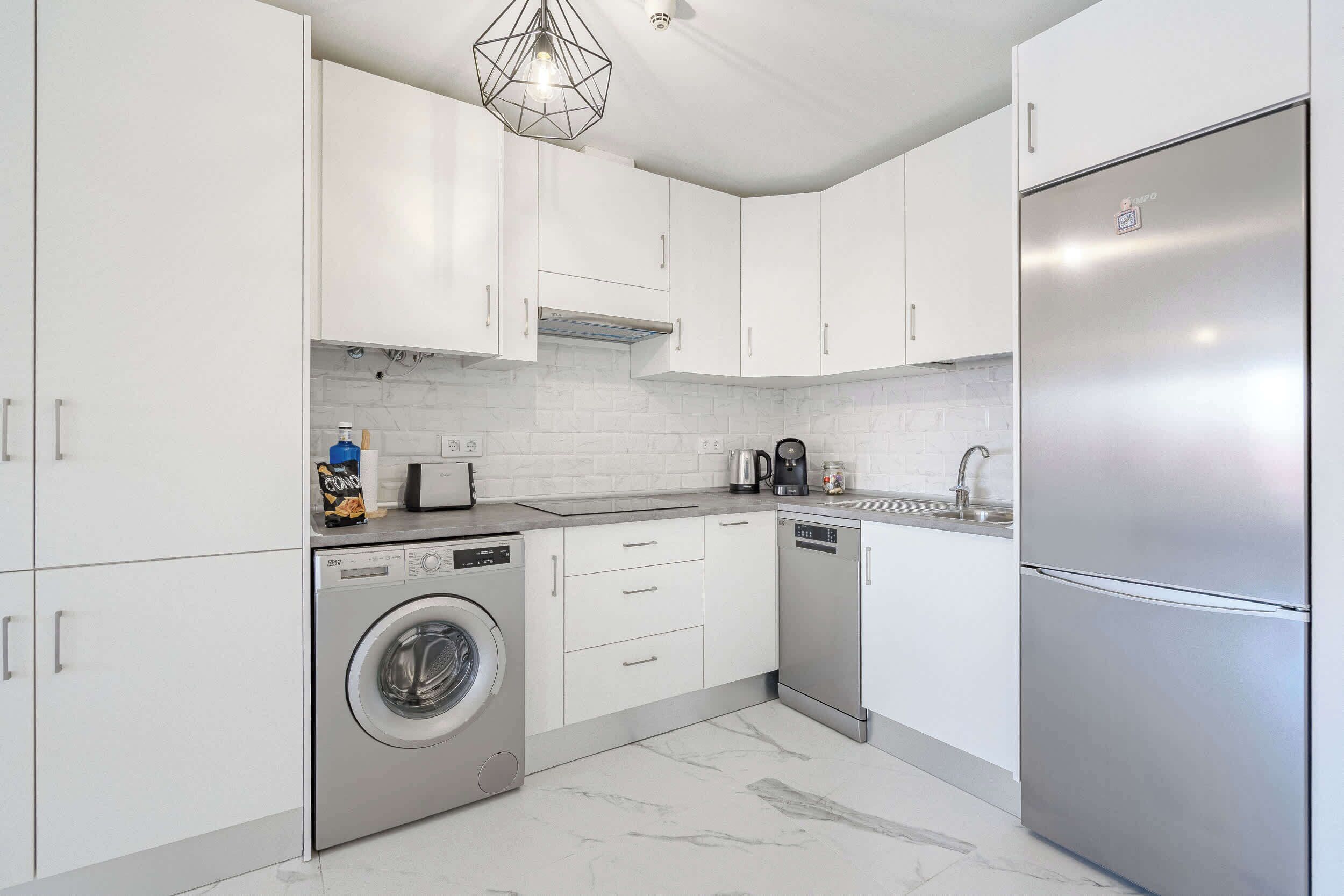 A modern kitchen with white cabinets, a stainless steel refrigerator, a washing machine, and a dishwasher.