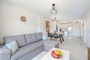 A modern living room with a grey sectional sofa, a dining area with a white table, and a kitchen in the background.