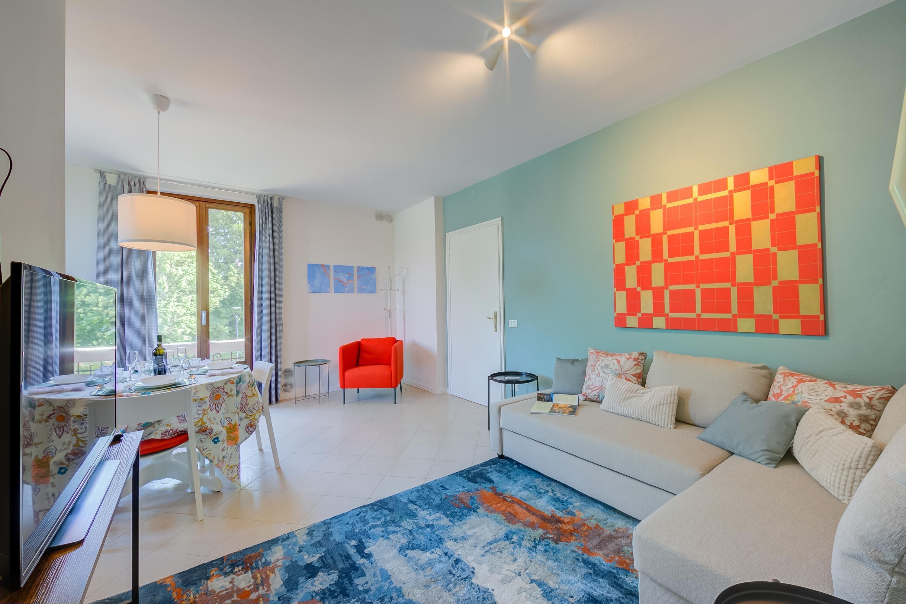 A modern living room with a sofa, a dining table, and a red chair. There is a large abstract painting on the wall and a window with curtains.