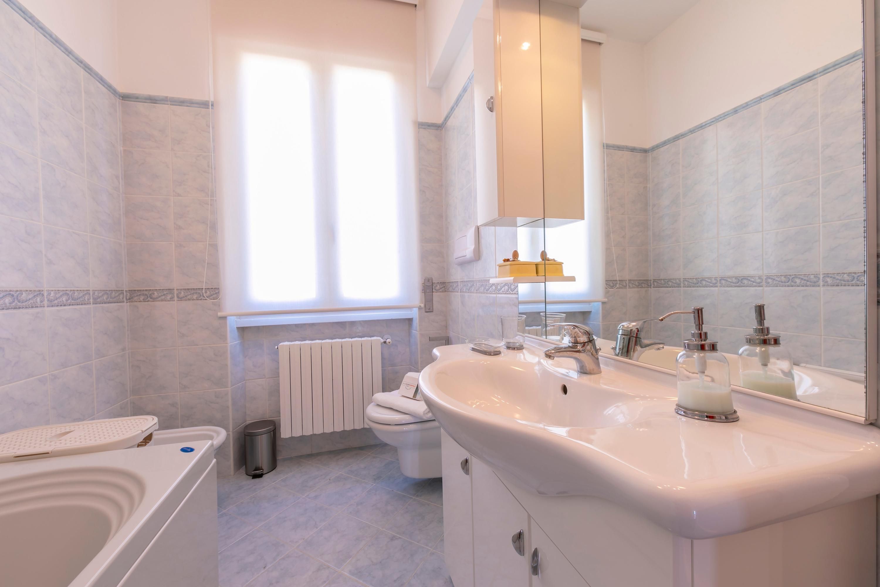 Une salle de bain moderne équipée d’une baignoire, d’un grand lavabo, de toilettes et d’une fenêtre laissant entrer la lumière naturelle.