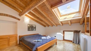A bedroom with a wooden ceiling, a skylight, a bed, a radiator, and a window with curtains.