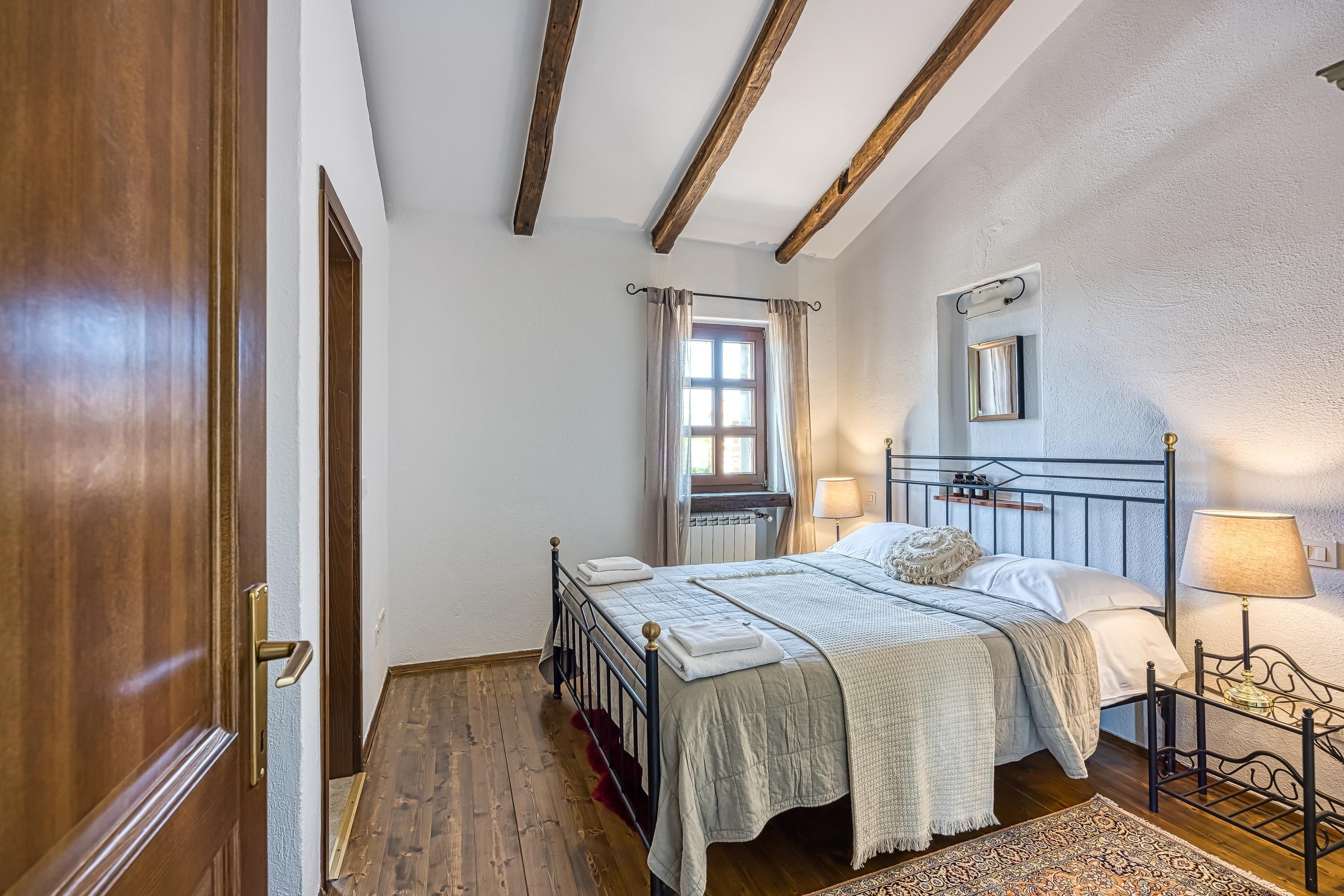 Une chambre avec un plancher en bois, un lit recouvert d’une courtepointe blanche, une table de chevet avec une lampe et une fenêtre avec des rideaux.