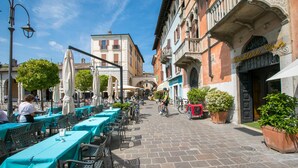 Un café al aire libre con mesas y sillas, un ciclista, y edificios con balcones y señalización.