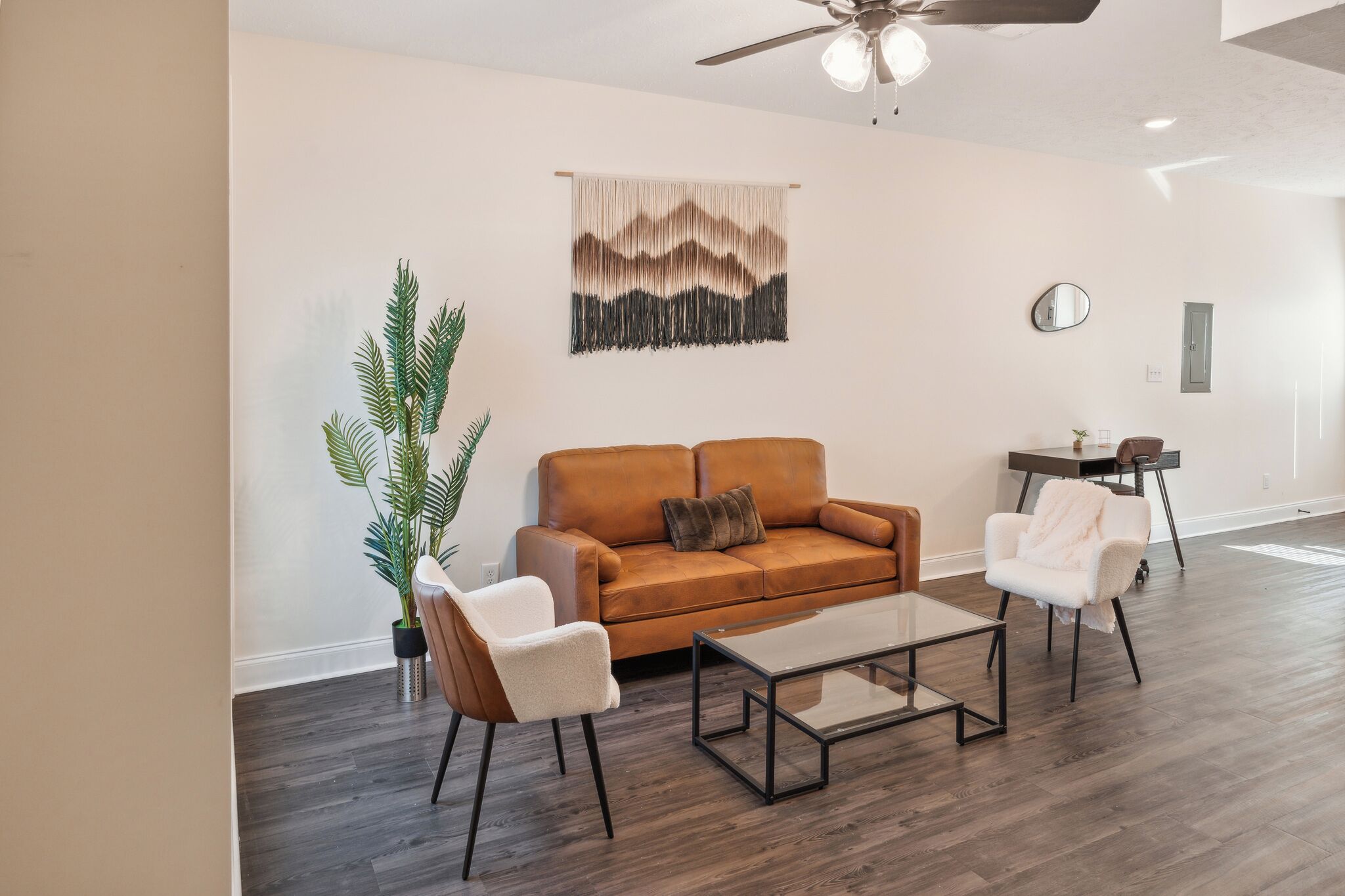 A modern living room with a brown leather sofa, a glass coffee table, and two white armchairs. A large plant is in the corner, and a wall tapestry is above the sofa.