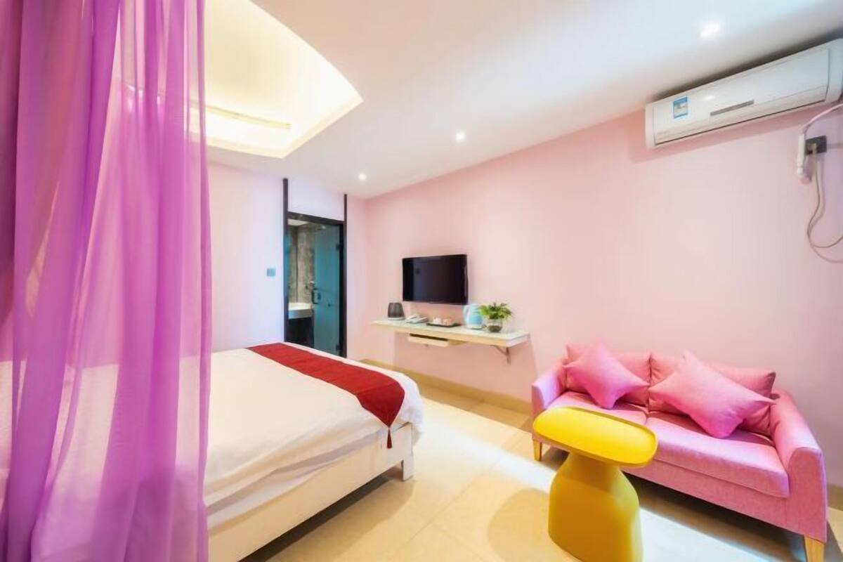 A bedroom with a bed, a TV, a pink sofa, and a yellow stool.