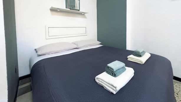 A neatly made bed with two folded towels, a bedside table with a mirror, and a wall-mounted shelf. - [Cactus-Room] (Padova)