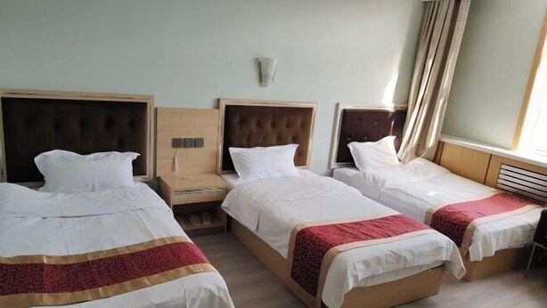 Une chambre d’hôtel avec trois lits simples, chacun doté d’une tête de lit en bois, de draps blancs et d’une couverture à rayures rouges et dorées.