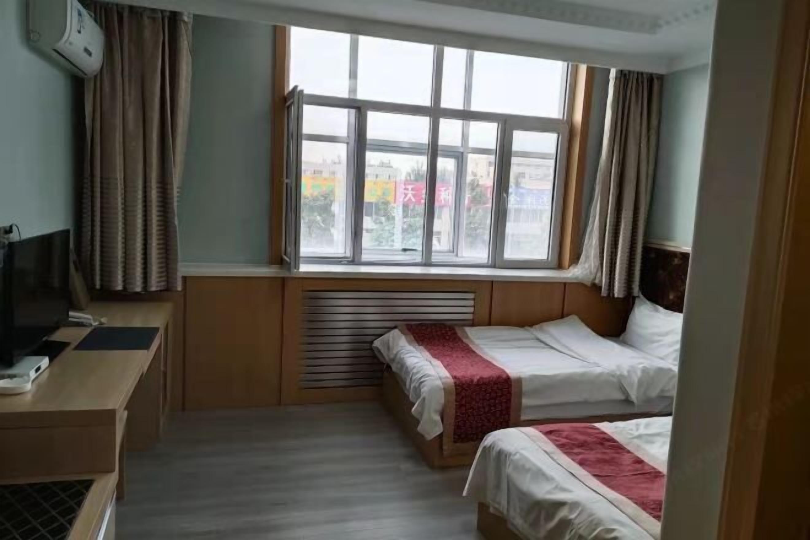 A hotel room with two beds, a desk with a computer, a window with curtains, and a wall-mounted air conditioner.