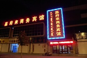 Ein Gebäude mit einer großen, beleuchteten Schrifttafel in chinesischen Schriftzeichen, ein Laden mit rotem Markisen und ein Baum davor.