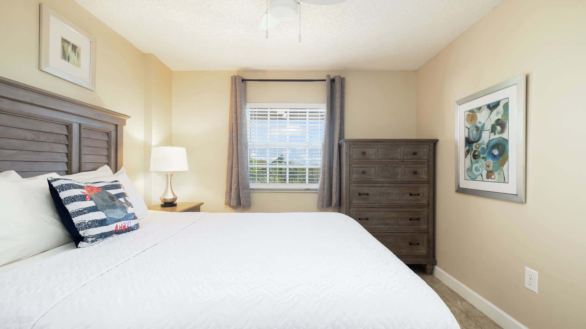 A bedroom with a bed, a nightstand, a dresser, a window with curtains, and two framed pictures on the walls.