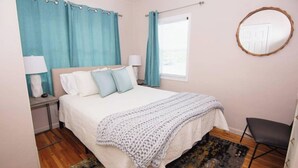 Ein Schlafzimmer mit einem Bett, zwei Nachttischen, einer Lampe, einem Fenster mit Vorhängen, einem Spiegel und einem Sessel.