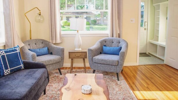 Ein Wohnzimmer mit zwei Sesseln, einem Sofa, einem Holztisch, einer Lampe und Blick auf ein Haus durch ein Fenster.