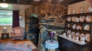 Ein gemütliches Wohnzimmer mit Kamin aus Stein, einer bequemen Couch, einem kleinen Tisch mit Lampe und einer Vitrine mit dekorativen Tellern.