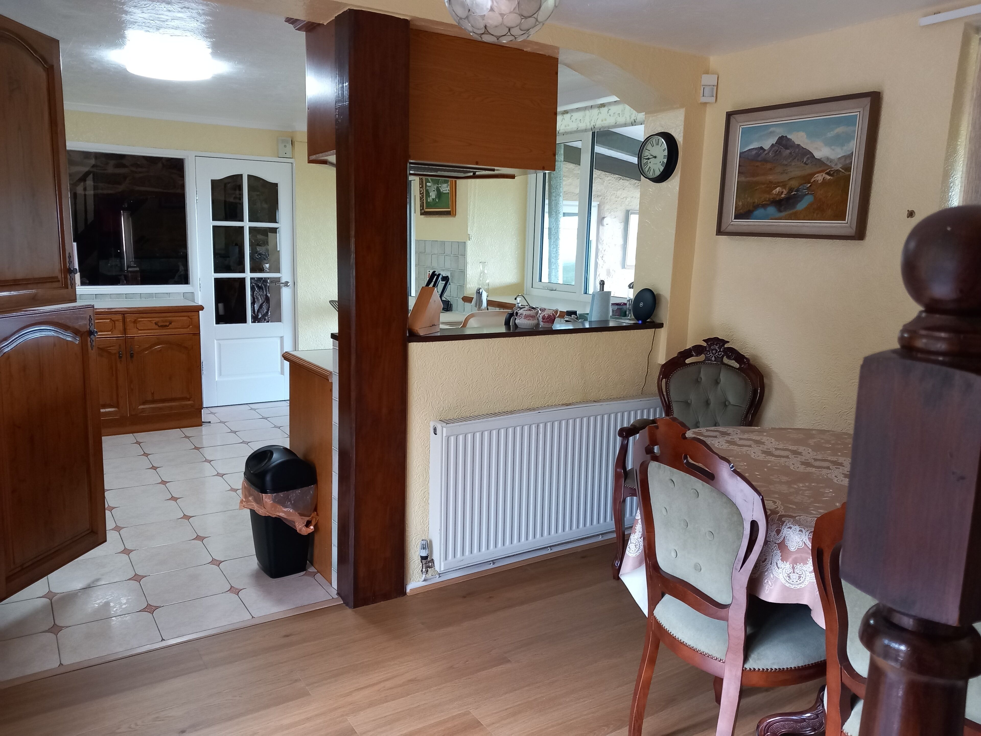 A dining area with a wooden table and chairs, a kitchen with wooden cabinets, a radiator, and a clock on the wall.