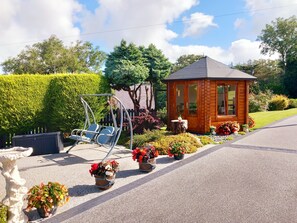 A wooden cabin with a small porch, surrounded by a garden with various potted plants and a swing chair. - Fox's Lair Farmhouse for Panoramic Views (Caernarfon)