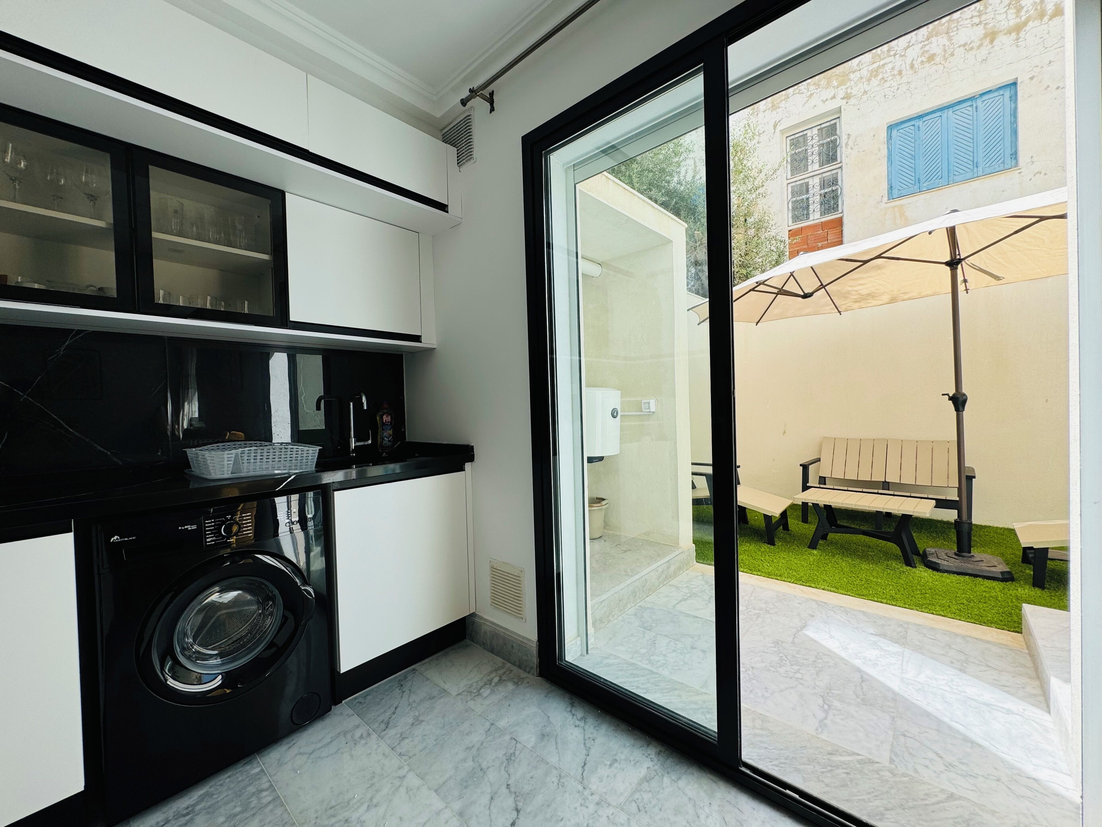 Een moderne keuken met een ingebouwde wasmachine en droger, een glazen deur naar een terras met een bank en parasol.