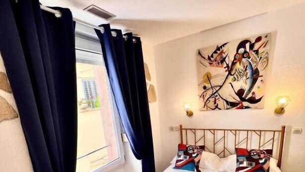 A bedroom with a bed, a window with curtains, a painting on the wall, and a laptop on the bed. - Hotel Appia (Cannes)
