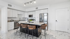 A modern kitchen with white cabinets, a marble island, bar stools, stainless steel appliances, and a large refrigerator.