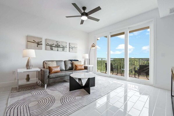 A spacious living room with a grey sofa, a coffee table, and a ceiling fan. There are large windows offering a view of the outdoors.